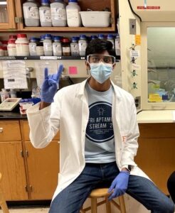 Mahit Vunnam pictured in a lab seated in front of lab equipment and wearing a lab coat, mask, and gloves while making the Longhorn sign with his right hand